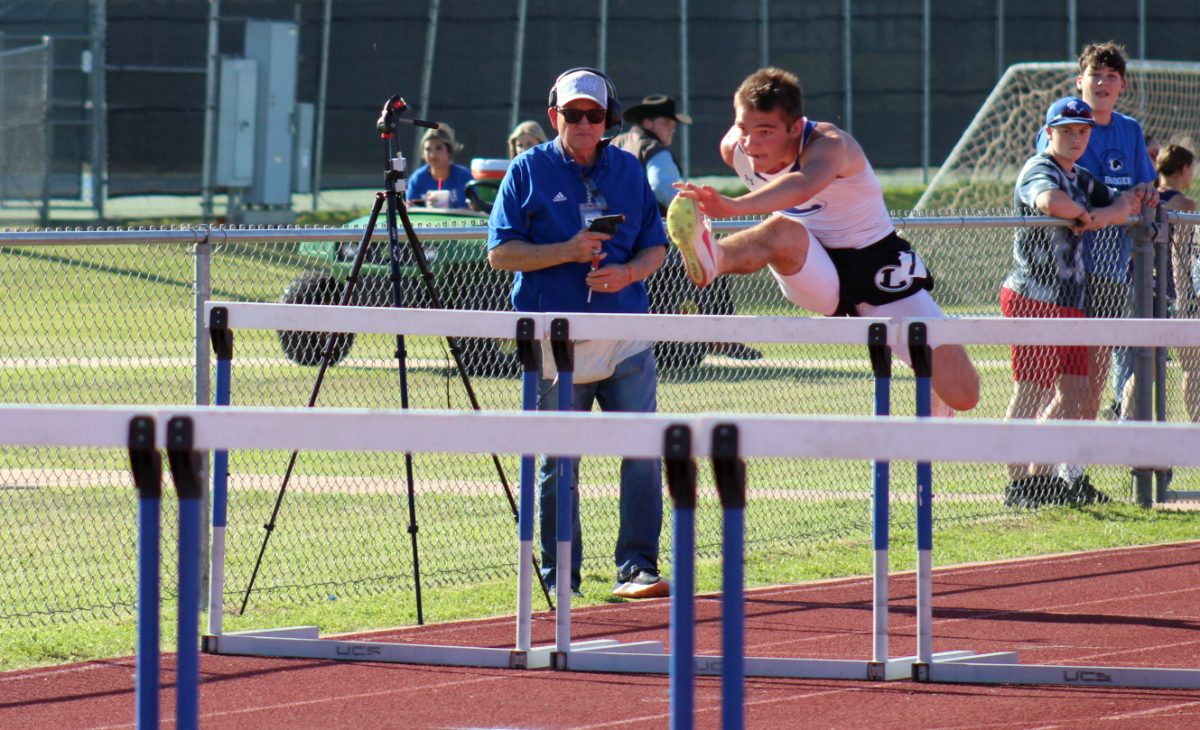 Sophomore Shawn Roberts hurdles during a race at the home track meet while Principal Joey McQueen holds the starting pistol. 